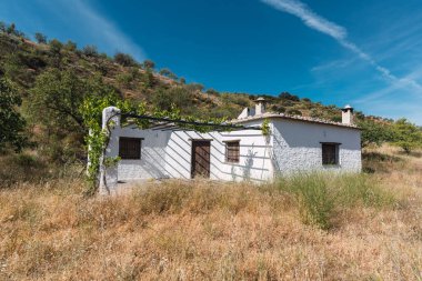 Kırsalda beyaz cepheli bir çiftlik evi, evin pencereleri ve parmaklıklı ahşap kapıları var, etrafında ağaçlar, gökyüzünde bulutlar var.