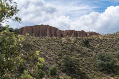 Dağ yamacında güçlü erozyonlarla dolu dağlık bir bölge. Çalı ve ağaçların bol bitkileri var. Gökyüzü bulutlu.