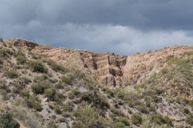 Dağ yamacında güçlü erozyonlarla dolu dağlık bir bölge. Çalı ve ağaçların bol bitkileri var. Gökyüzü bulutlu.
