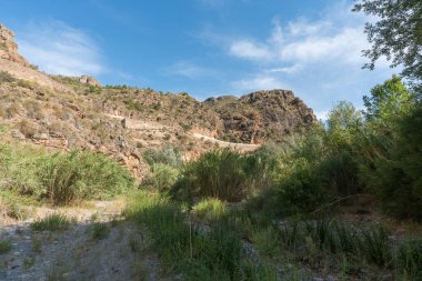 Güney İspanya 'da dağlık bir arazi, bir nehir var ve kenarında kamış tarlaları ve kavak ağaçları var, dağlarda çalılar var, gökyüzü çok az bulutla açık.