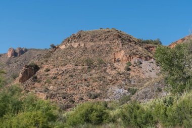 Güney İspanya 'nın dağlık arazisi, kamış tarlaları ve ağaçları var, dağda büyük kayalar var, gökyüzü açık.