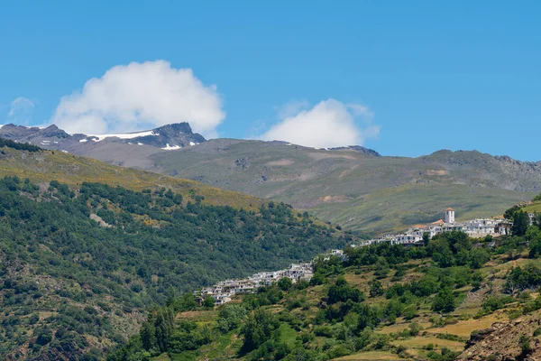 The town of Capileira in the Sierra Nevada mountains, the mountains have pine forest and shrubs, the mountains have snow, the sky has clouds