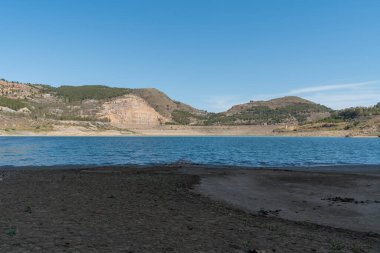 Beninar rezervuarı dağlarla çevrili, dağlarda ağaçlar ve çalılar var, gökyüzü açık.