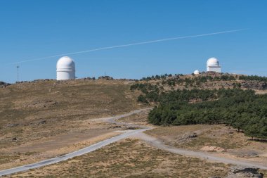 Calar Alto Astronomik Gözlemevi, tesise giden bir yol var, çam ormanı var, kayalar ve bitki örtüsü var ve gökyüzü açık.