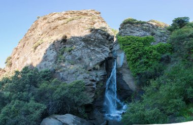 Sierra Nevada 'da bir nehirde şelale, bir kaya duvarı, kayalar, ağaçlar ve çalılar var, gökyüzü açık.