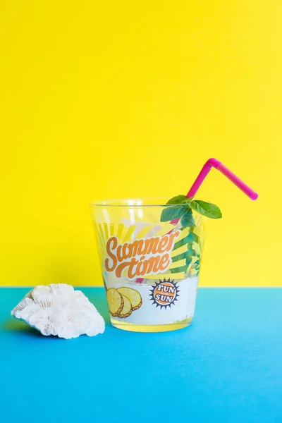 A glass of refreshing drink with straw, aromatic basil leaves and sea shells on a yellow and light  blue background. With stamped summertime and fun in the sun. Different views