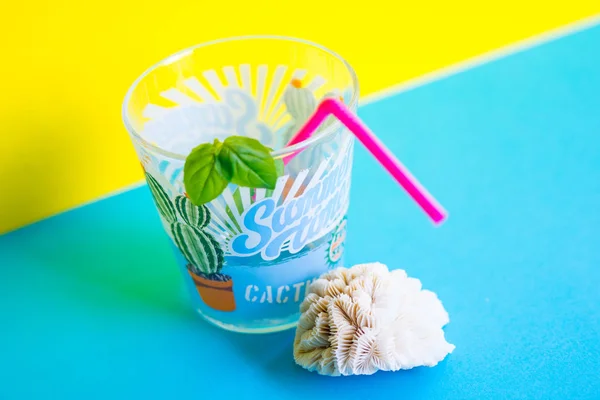 Close-up of a glass of refreshing drink with straw, aromatic basil leaves and sea coral on a yellow and light  blue background. With stamped summertime and fun in the sun. Different views.
