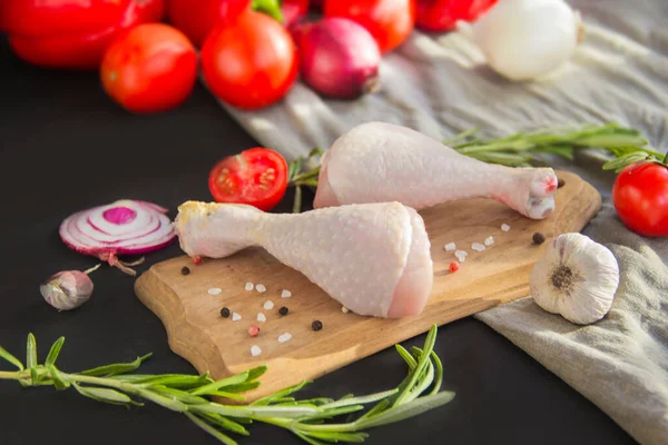 Raw chicken legs with rosemary, tomatoes, and spices on a black wooden background. Creative atmospheric decoration.