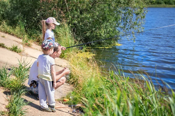 Daughter with father and brother sit on the shore of the lake with rods and catch fish. Concept of outdoor recreation and holiday with family and fishing . Father teaches his children to catch a fish.