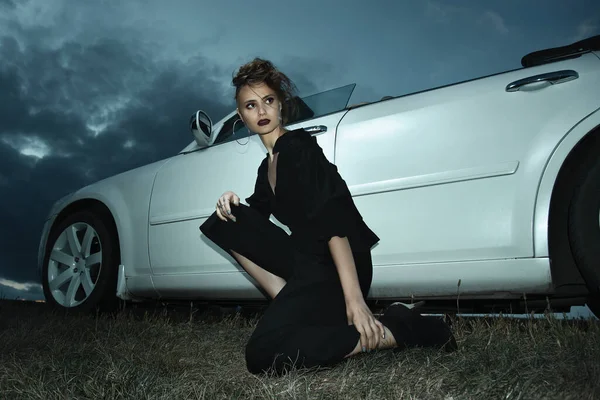 Şık kız gün batımında üstü açık beyaz bir arabanın yanında poz veriyor.