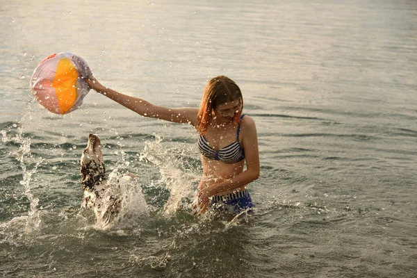Νεαρή ελκυστική κοπέλα παίζει μπάλα με το σκυλί στο μπλε νερό της λίμνης — Φωτογραφία Αρχείου