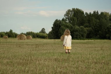 Köyün tarzında giyinmiş bir kız samanlı bir tarlada duruyor. Arkadan görüntüle.