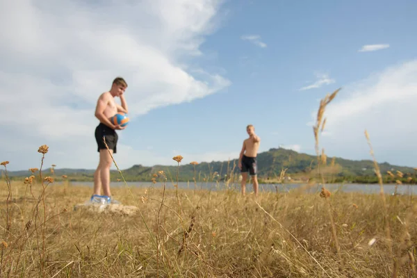 Dwóch gości gra w siatkówkę na tle górskiego jeziora. Koncentracja na trawie — Zdjęcie stockowe