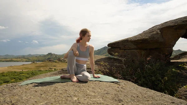 Une fille en costume de yoga gris est assise sur un rocher. Repos pendant l'entraînement. Entraînement sportif sur fond de paysage montagneux. Images De Stock Libres De Droits