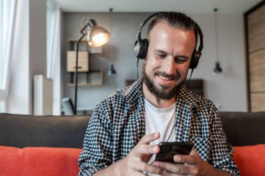 Adam kulaklıkla müzik dinliyor, kanepeye oturuyor ve akıllı telefonuna bir mesaj yazıyor.