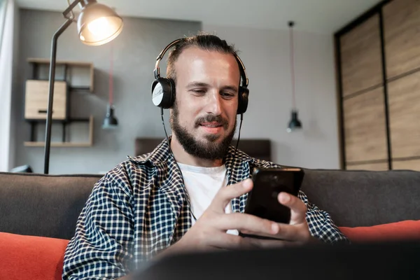 The guy listens to music on headphones, sits on the couch and writes a message on his smartphone
