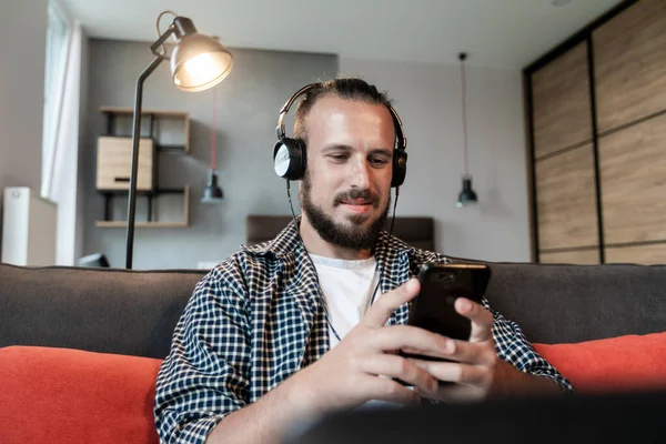 The guy listens to music on headphones, sits on the couch and writes a message on his smartphone