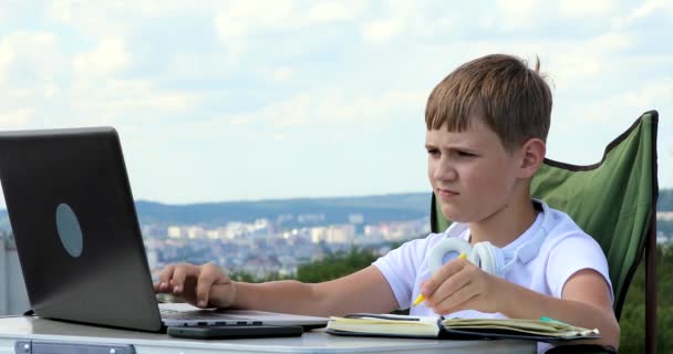 那个戴耳机的男孩在电脑前学习 在笔记本上做笔记 结果证明一切都很好 — 图库视频影像