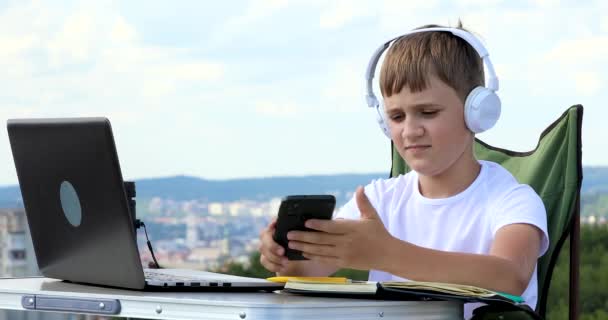 Pojke Hörlurar Tittar Telefonen Och Gör Anteckning Laptop — Stockvideo
