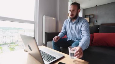 Kulaklıklı genç Amerikalı adam kanepede yatar ve dizüstü bilgisayarına yazar ve telefonda konuşur.