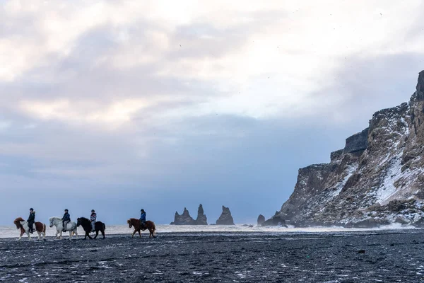 İnsanlar, Reynisfjara 'daki Atlantik Okyanusu' nun dalgaları ve panoramasının kara sahillerinde atlarla uzaklaşıyorlar.