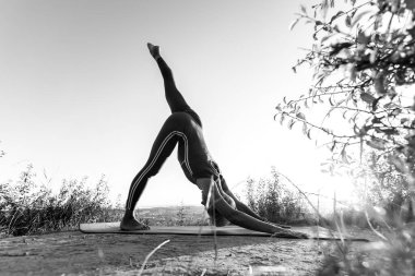 Siyah ve beyaz yaşlı kadın yoga poziyonu veriyor. Güneşin doğuşuyla ilgili bir arka planda baş aşağı köpek pozisyonu alıyor.
