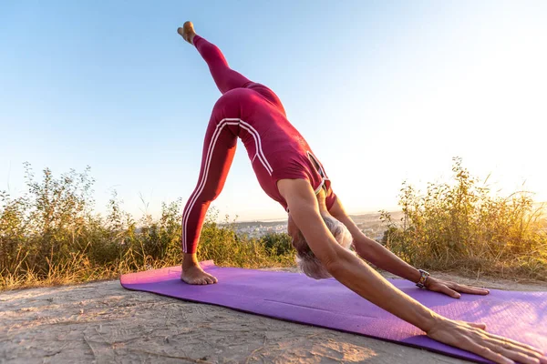 Önden bakan yaşlı kadın yoga pozisyonu alıyor. Başını kuma yaslamış köpek pozisyonu alıyor.