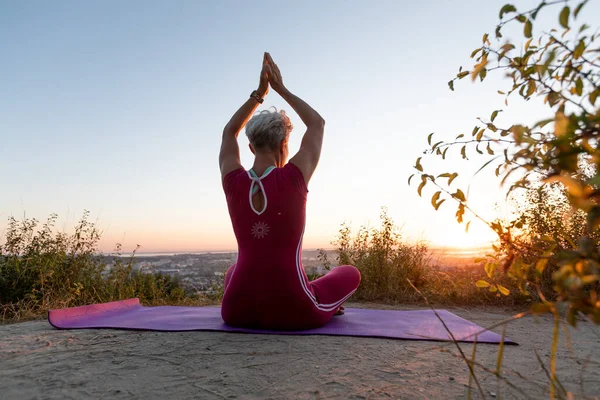 Woman back view holding hands up while sitting in lotus position on morning sunrise and town sky background