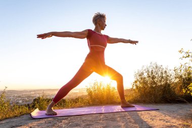 Kırmızı pantolonlu yaşlı kadın yoga pozisyonu yapıyor. Savaşçı Virabhadrasana 'yı Aurora' da keşfet.