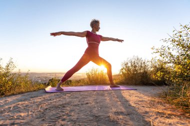 Güneş doğarken sana pozisyonunu veren kadın Virabhadrasana II kırmızı pantolonlu.