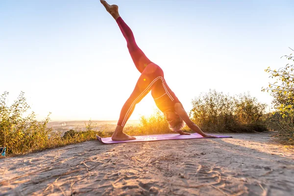 Kırmızı pantolonlu kadın yoga poziyonu alıyor. Baş aşağı köpek gün doğumunda bacağını kaldırdı.