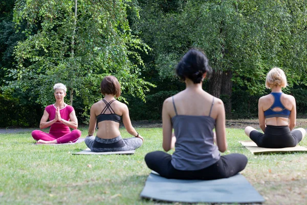 Female yoga master sits on a sports mat and teaches a group of girls in a lotus position and holds palms together outdoors on the ground in the park