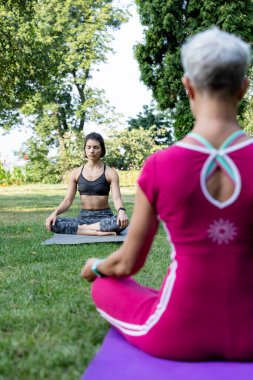 Kız çimlerin üzerinde lotus pozisyonunda oturuyor ve yoga ustasının rehberliğinde dinleniyor.