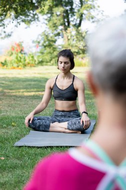 Güzel bir kız çimlerin üzerinde lotus pozisyonunda oturur ve yoga ustasının rehberliğinde kendini sakin hisseder.