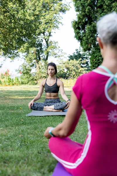 Kız çimenlerin üzerinde lotus pozisyonunda oturur ve yoga ustasının rehberliğinde kendini huzurlu hisseder.