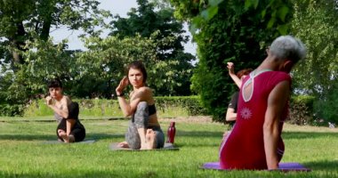 Bir grup sporcu yoga antrenörü eşliğinde çim parkında yoga yapıyor.