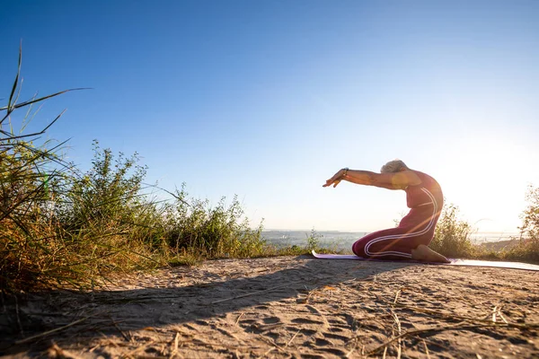 Kadın kumda oturuyor ve yoga pozunda kollarını öne doğru açıyor.