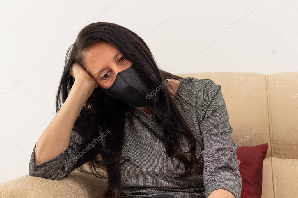 Middle-aged woman wearing face mask seated in armchair with a hand holding her head and thinking. Looks confused and lonely.
