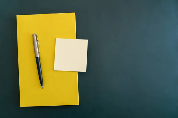 Yellow book with pen and sticky notes on dark green flat background.