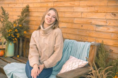 Sonbahar havası. Kahverengi süveter ve kot pantolon giymiş gülümseyen bir kız ahşap bir duvara karşı bankta oturuyor.