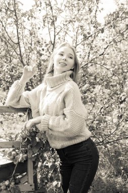 Rahat bir kazak ve kot pantolon giymiş gülümseyen bir kız bahçede yürüyor ve elma topluyor. Monokrom sürümü.