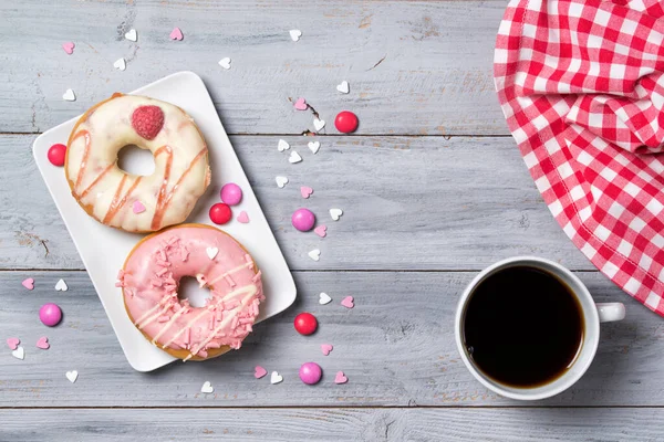 İki pembe donut, süslü püslü kalpler ve bir fincan kahve, ahşap arka plan.