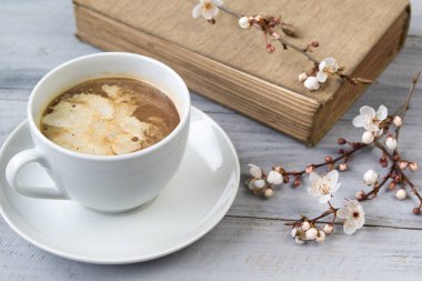 Kremalı bir fincan kahve ve ahşap zemin üzerinde çiçek açan kiraz dalları olan eski bir kitap.