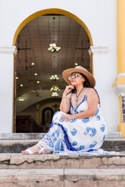 Mavi beyaz elbiseli bir kadın kilisenin merdivenlerinde oturuyor, hasır şapka ve güneş gözlüğü takıyor..