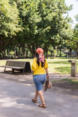 Sarı tişörtlü ve turuncu başörtülü bir kadın elinde hasır şapkayla parkta yürüyor.