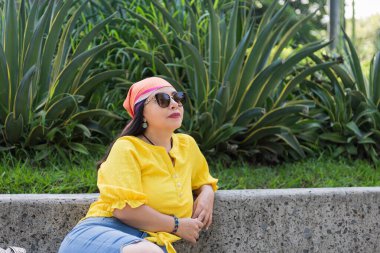 Sarı bluz ve başörtüsü takmış bir kadın açık havada, güneş gözlüklü, yemyeşil bitkilerin yanında dinleniyor..