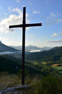 Hıristiyan haçı bir dağda, sabahın erken saatlerinde, mavi gökyüzü, vadide sis, arkada dağlar, La Beaume, Hautes Alpes, Fransa