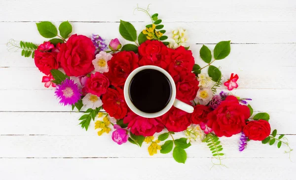 floral summer composition of roses, peonies, cornflowers, green leaves and a Cup of black coffee on a white wooden background, top view flat lay