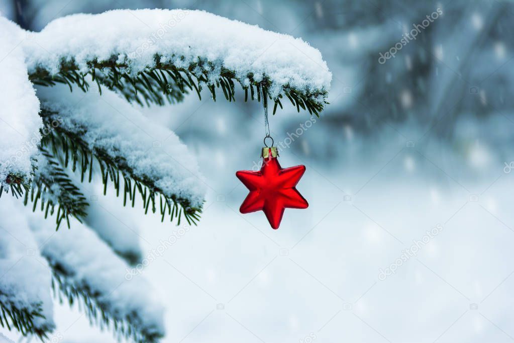 red Christmas tree toy in the shape of a star on a snow-covered branch of the Christmas tree and falling snowflakes on a cold winter day