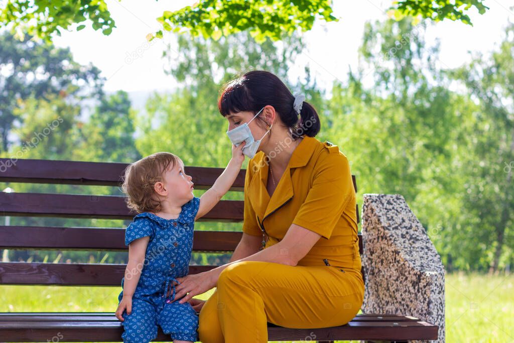 a young mother with a small baby daughter in a summer Park, a girl tries to take off  the medical mask from her mother, the concept of ending the coronavirus pandemic